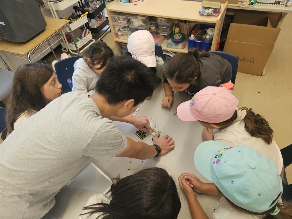 A mentor wiring a breadboard while elementary students crowd around the table at Dublin Heights